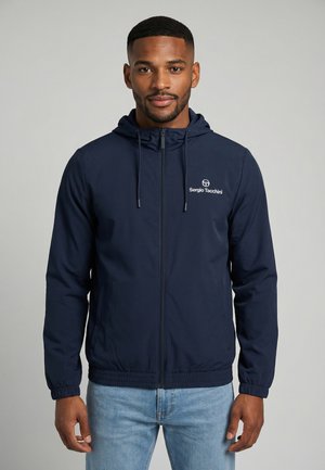 Hombre que lleva una chaqueta con capucha y cremallera de color azul marino de Sergio Tacchini y vaqueros azul claro, de pie frente a un fondo gris liso.