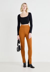 Zwart langemouwen cropped top met een textuurpatroon; ribbelige oranje hoge taillebroek met zijopeningen; model houdt een kleine zwarte tas vast.