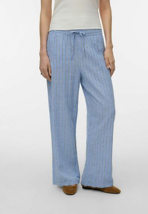 Mujer con pantalones holgados de color azul claro con rayas verticales blancas y azul marino, combinados con zapatos marrones y una blusa blanca.