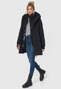 Schwarze gepolsterte Jacke mit Kapuze, Reißverschluss- und Knopfverschluss sowie strukturiertem Stoff. Getragen über einem grauen Oberteil, kombiniert mit blauen Jeans und schwarzen Ankle Boots.