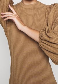 Vestido marrón texturizado con cuello alto y mangas abullonadas. El tejido presenta un patrón de cuadrícula y detalles fruncidos en los puños de las mangas.