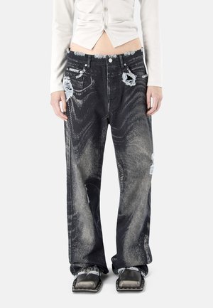 Persona que lleva pantalones vaqueros negros desgastados y holgados con patrones descoloridos, combinados con zapatos negros de punta cuadrada y una blusa blanca corta con botones.