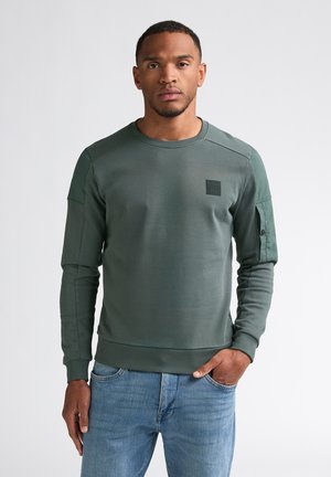 Man met kort haar die een groene longsleeve trui en een blauwe spijkerbroek draagt, staand met één hand in zijn zak tegen een effen achtergrond.