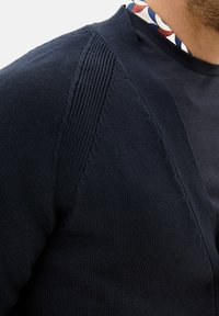 Marinblå kavaj med ribbad textur längs axelsömmarna, med en underliggande t-shirt med rund halsringning som har ett flerfärgat kragmönster.