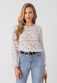 White lace top with floral patterns and long sleeves, paired with high-waisted blue jeans and a black belt, featuring ruffled sleeve edges.