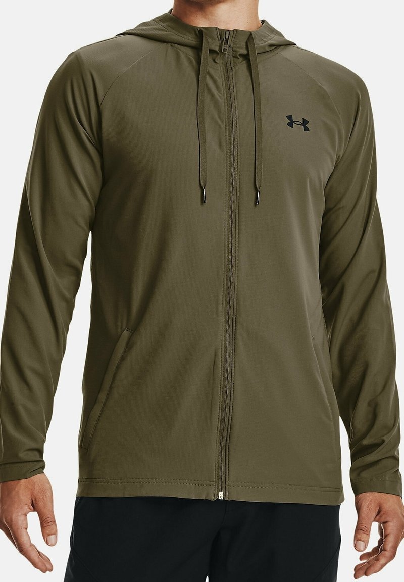 Homme portant un sweat à capuche zippé vert olive avec cordons et logo Under Armour, associé à un pantalon noir, visible du cou jusqu’aux hanches.