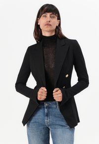 Zwarte dubbelgeknoopte blazer met gouden knopen, op maat gemaakt ontwerp en lange mouwen, gedragen over een aangesloten zwart topje en blauwe denim jeans.