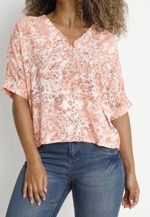 Kvinde iført en løs, kortærmet lyserød og hvid mønstret skjorte med knapper og blå jeans, stående foran en ensfarvet baggrund.