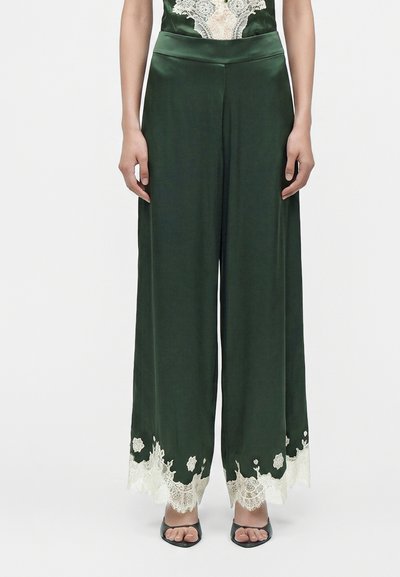 Femme portant un pantalon large vert foncé avec une bordure en dentelle florale blanche à l’ourlet et des talons ouverts noirs.