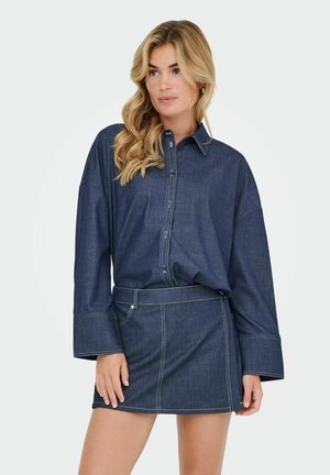 Femme aux longs cheveux blonds portant une chemise en jean bleu foncé à boutons et une mini-jupe en jean assortie, se tenant avec sa main reposant sur sa cuisse.