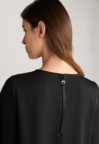 Zwarte jurk met een ronde halslijn, gladde textuur en een rits aan de achterkant met een kleine metalen trekker. Gestyled met minimale naden.