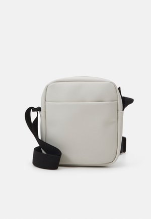 Petit sac bandoulière beige avec une sangle noire réglable, de forme carrée, et une poche avant à rabat sur un fond blanc.