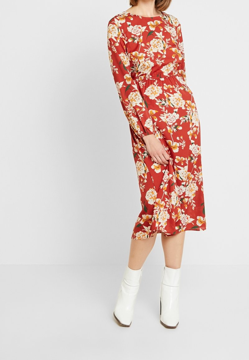 Femme portant une robe midi rouge fleurie à manches longues associée à des bottines blanches, debout devant un fond clair uni.