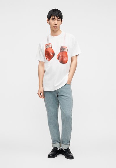 Λευκό βαμβακερό T-shirt με μεγάλο γραφικό κόκκινων γαντιών πυγμαχίας με το κείμενο "MOSCHINO". Συνδυάζεται με ανοιχτόχρωμα μπλε τζιν και μαύρα παπούτσια.