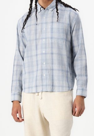 Mężczyzna ubrany w jasnoniebieską koszulę w kratę z długim rękawem na guziki oraz kremowe spodnie, stojący z rękami swobodnie opuszczonymi wzdłuż ciała.