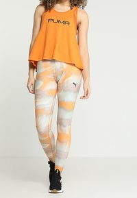 Top senza maniche arancione con logo "PUMA" nero, abbinato a leggings fantasia in arancione, grigio e bianco. Sneakers nere con dettagli dorati.