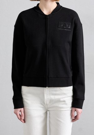 Osoba nosząca czarną kurtkę na zamek z logo EA7 Emporio Armani na piersi i białe spodnie, stojąca na jednolitym tle.