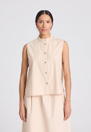 Kvinde med mørkt hår iført en ærmeløs beige skjorte med knapper og matchende nederdel, stående mod en ensfarvet lys baggrund.