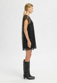 Moteris su šviesiais plaukais surištais į uodegą, vilkinti juodą be rankovių suknelę su tinklelio raštu ir juodais iki kelių batais, stovinti profiliu.