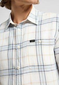 Camisa de cuadros clara en tonos blanco, azul y beige. Cuenta con cuello abotonado, un bolsillo frontal y una etiqueta negra con el logo "Lee".