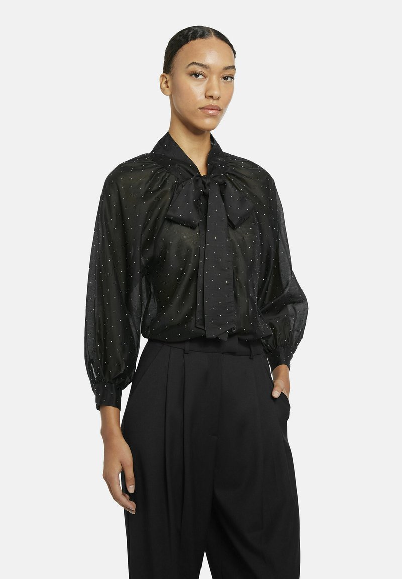 Black sheer blouse with large bow tie at neckline, puffy sleeves, and white polka dot pattern. Paired with high-waisted black trousers.