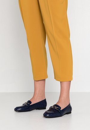 Navy blue loafers with a textured crocodile pattern, featuring a decorative silver chain accent and a flat sole. Paired with mustard-colored trousers.