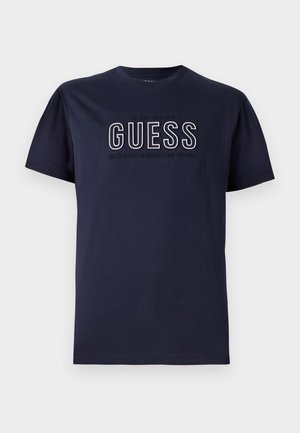 T-shirt in cotone blu navy con maniche corte, caratterizzato da un logo in rilievo "GUESS" e la scritta "CLASSIC AMERICAN STYLE" in bianco e grigio. Design semplice.