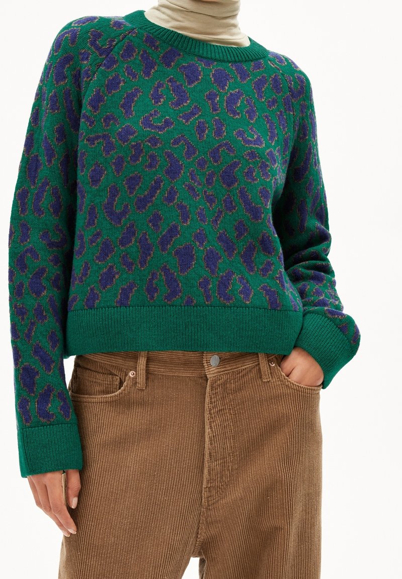 Pull vert et violet à imprimé léopard porté sur un col roulé beige, associé à un pantalon en velours côtelé marron taille haute, main dans la poche.
