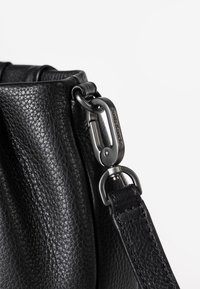 Schwarze Lederhandtasche mit strukturierter Oberfläche, verstellbarem Trageriemen und einem metallischen Verschluss mit graviertem Markenlogo.