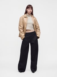 Femme portant une veste courte beige, un crop top à col roulé rayé et un pantalon cargo large noir, debout sur fond blanc.