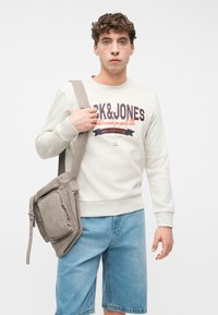 Νέος άνδρας που φοράει ανοιχτόχρωμο φούτερ με το κείμενο «JACK&JONES», τζιν σορτς και κρατάει μια μπεζ τσάντα χιαστί στον ώμο, κοιτάζοντας μπροστά.