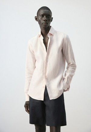 Męski model ubrany w jasnoróżową, długorękawową koszulę na guziki oraz czarne szorty, stojący na jednolitym, jasnym tle.