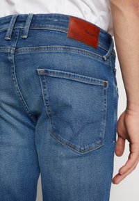 Mörkblå denimjeans med rak passform, med en bältepatch i brunt läder och kontrasterande sömmar. Bakfickan har en böjd design.