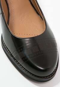 Ballerines en cuir noir avec un bout arrondi, présentant des motifs texturés et un bord cousu. La semelle intérieure est de couleur marron clair.