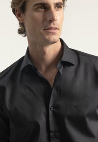 Camisa negra de botones fabricada en un tejido suave, con cuello puntiagudo, logotipo sutil en el pecho y botones negros.