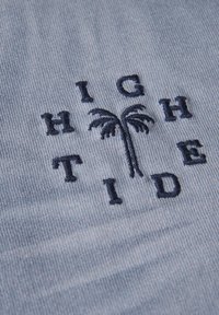 Světle modrá látka s námořnickým vyšíváním, které tvoří nápis "HIGH TIDE" spolu s ikonou palmy. Textura je měkká s hladkým, mírně žebrovaným povrchem.