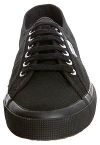 Svart canvas sneakers med en texturerad yta, vita öljetter och runda snören. Har en svart gummisula med ett mönstrat grepp.