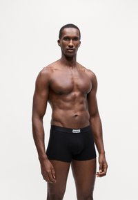 Sorte bomullsboxershorts med merket midjebånd. Modellen står oppreist og viser frem passformen og designet. Enkelt mønster.
