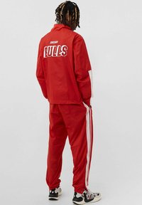 Tuta rossa con accenti bianchi, con il logo "CHICAGO BULLS" sul retro. Realizzata in materiale liscio e leggero. Sneakers nere e bianche.