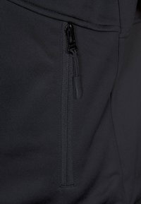 Czarny materiał sportowej odzieży z zamykaną na suwak boczną kieszenią, gładką fakturą i dopasowanym krojem. Suwak jest dyskretny i funkcjonalny.