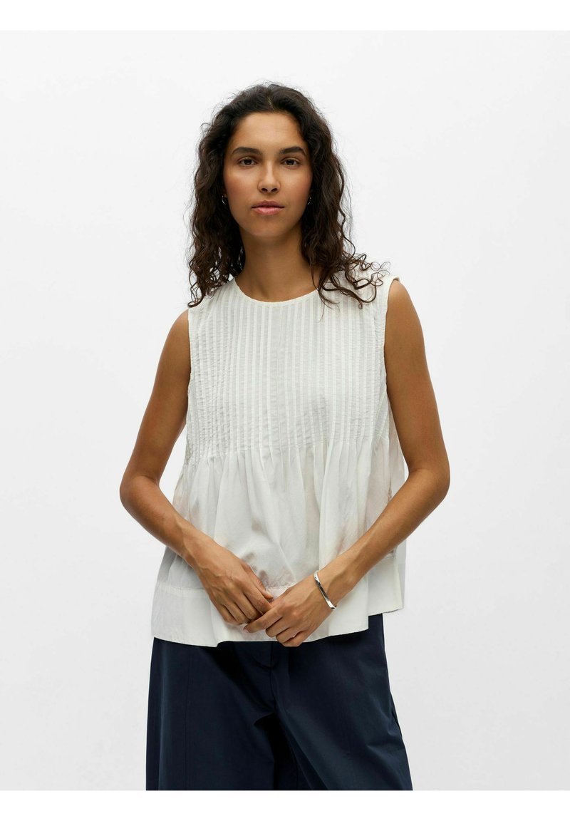 Femme aux cheveux bouclés portant un chemisier blanc texturé sans manches et un pantalon foncé, debout, les mains jointes de manière lâche devant elle.