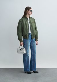 Žena v zelenej bomber bunde, bielej košeli, modrých džínsoch, čiernych lodičkách a slnečných okuliaroch, drží bielu kabelku s čiernym textom a grafikou.