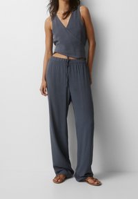 Grijze mouwloze wikkeltop met V-hals, gecombineerd met losse grijze broek met elastische tailleband en trekkoord. Platte sandalen maken de look compleet.