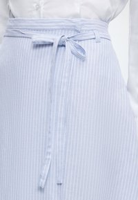 Φούστα με κάθετες ρίγες σε ανοιχτό γαλάζιο και λευκό, δεμένη με φιόγκο στη μέση, συνδυασμένη με λευκό πουκάμισο με κουμπιά.