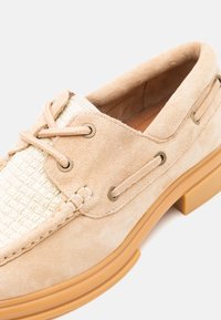 Beige semskesko med vevet kremfarget stoffinnlegg og runde snører. Har metalliske öyelets og en brun gummisåle.