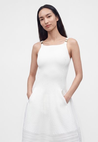 Femme aux longs cheveux foncés portant une robe blanche texturée sans manches avec des bretelles à boucle dorée et les mains dans les poches, sur un fond uni.