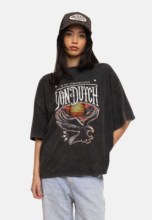 Jeune femme portant un t-shirt graphique noir Von Dutch avec un design d'aigle et un jean bleu clair, assorti à une casquette de baseball Von Dutch à motif.