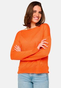Pull en tricot orange vif avec un large col bateau et des manches longues. Associé à un jean en denim bleu clair. Texture douce, design minimal.