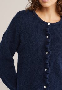 Tamsiai mėlynas mezgimo megztinis su minkšta tekstūra, puoštas raukšlėta sagų juosta ir apvalia iškirpte, su baltais apvaliais mygtukais.