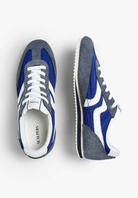 Zapatillas de ante azul y gris con acentos blancos, que presentan un diseño retro, suela de goma y cordones tradicionales. Marca 'Scalpers' visible en el interior.
